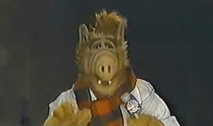 ALF Hosting Macy's Thanksgiving Day Parade - A furry alien character, reminiscent of ALF, wears a red and black checkered scarf and white sweater while holding an NBC microphone. With a snout-like nose, they appear to be hosting the Macy's Thanksgiving Day Parade.