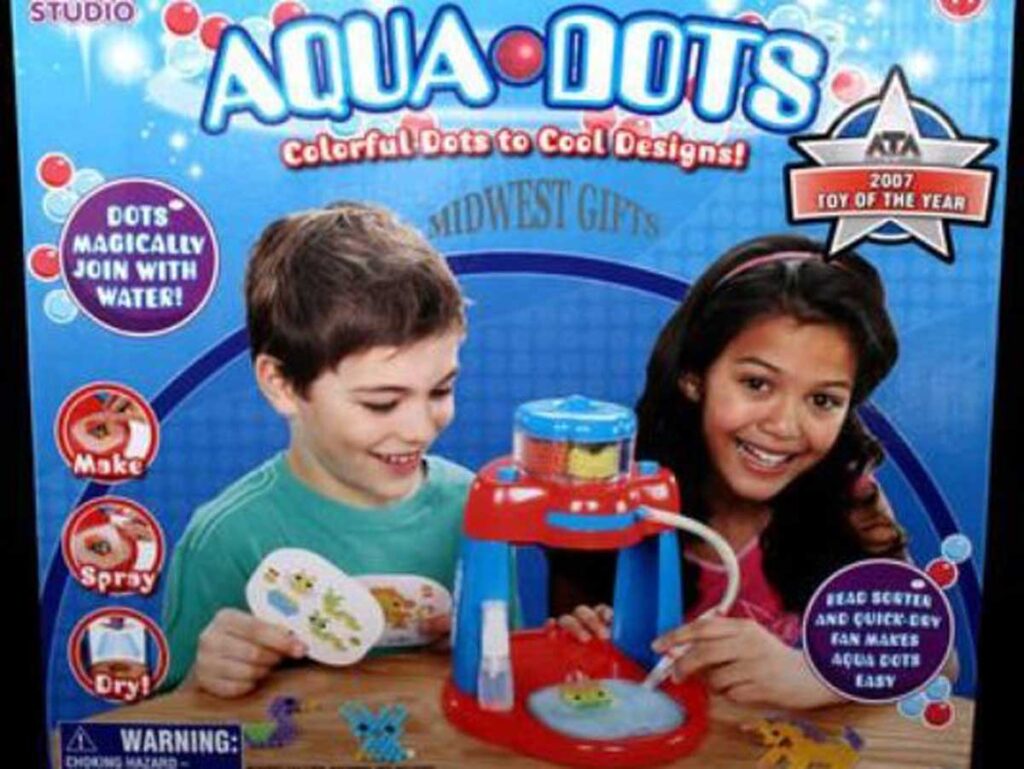 A box for Aqua Dots, a childrens craft toy, features two kids using the product. Text describes it as Colorful Dots to Cool Designs and 2007 Toy of the Year. The toy promises to magically join dots with water and includes a warning.