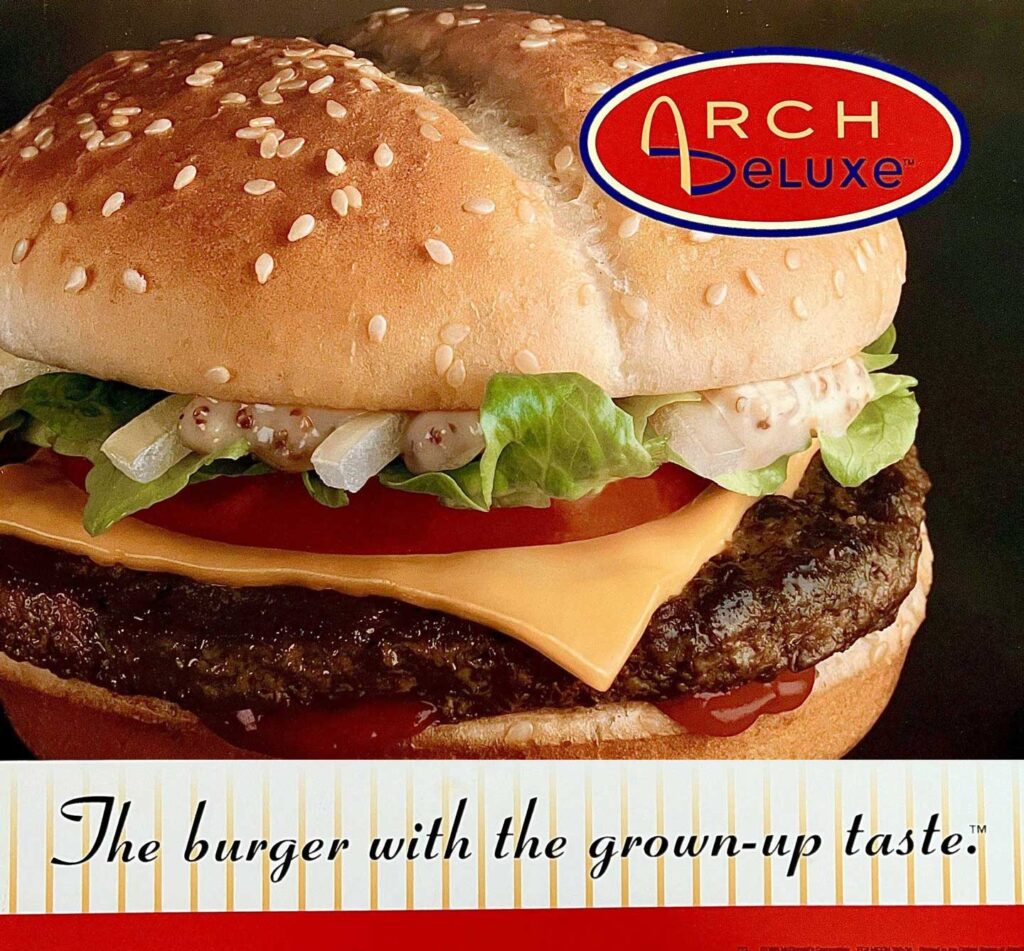 Discontinued fast food items - Arch Deluxe. A close-up of a burger featuring a sesame seed bun, lettuce, tomato, onion, a beef patty, and cheese. The Arch Deluxe logo is at the top right with "The burger with the grown-up taste" slogan below. A nostalgic nod to discontinued fast food items that left an indelible mark.