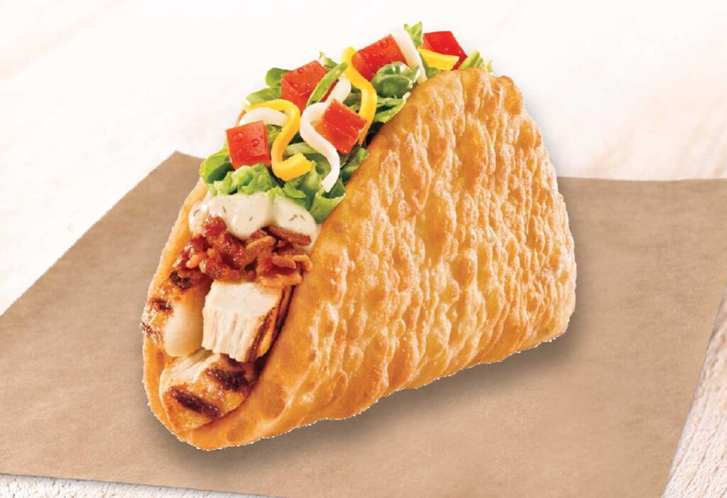 A crispy taco shell filled with grilled chicken pieces, lettuce, diced tomatoes, shredded cheese, and sauce rests on a brown paper surface—a nostalgic nod to discontinued Taco Bell menu items.