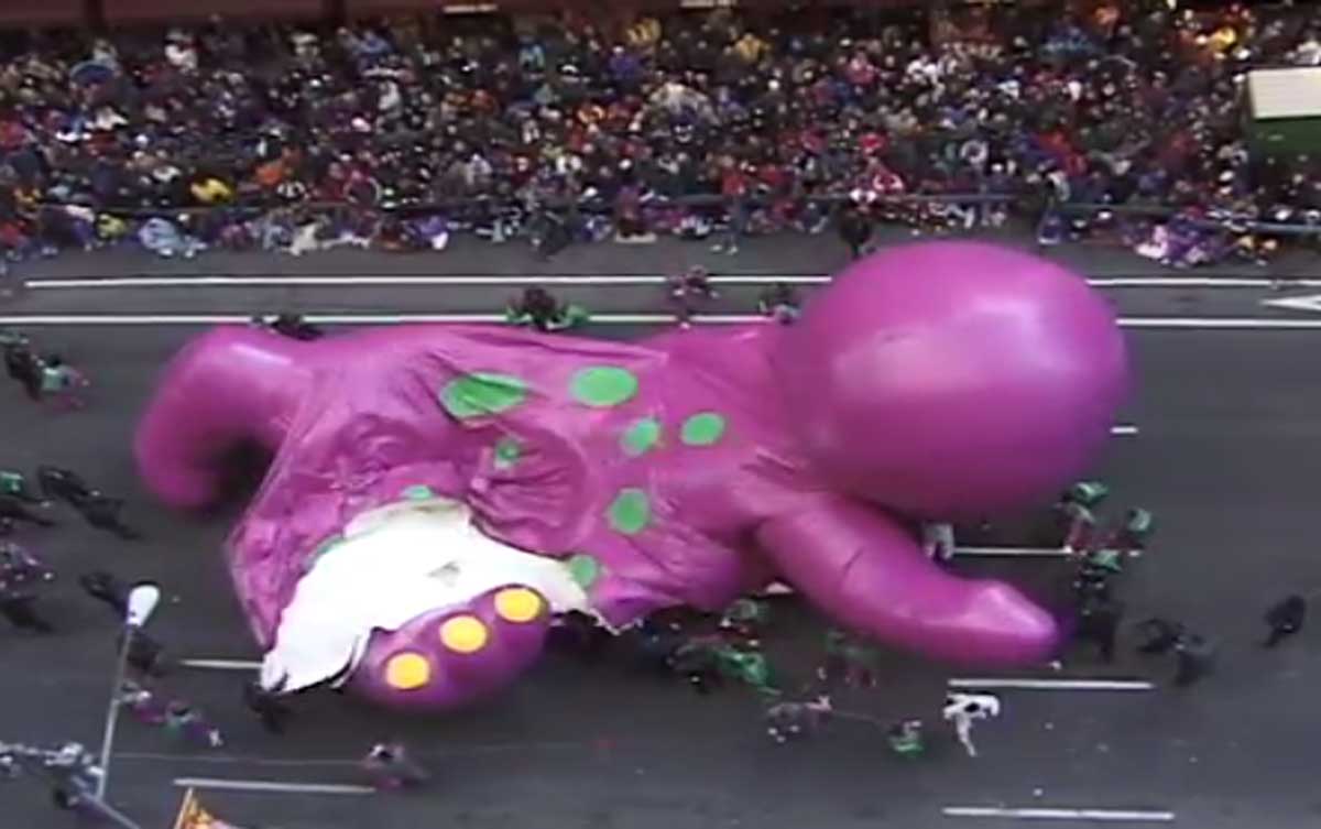 A large, deflated purple dinosaur balloon lies on the street during a parade. Several people surround it, attempting to manage the situation. A crowd watches the scene from behind barricades.