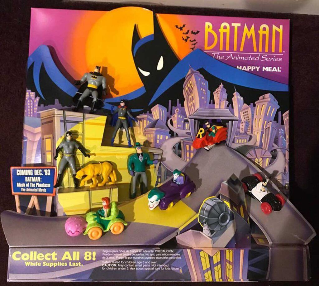 A vintage Batman: The Animated Series Happy Meal display features action figures and toys from the series with characters like Batman, Robin, and the Joker. The backdrop shows a stylized Gotham City with the Bat-Signal shining in the night sky.