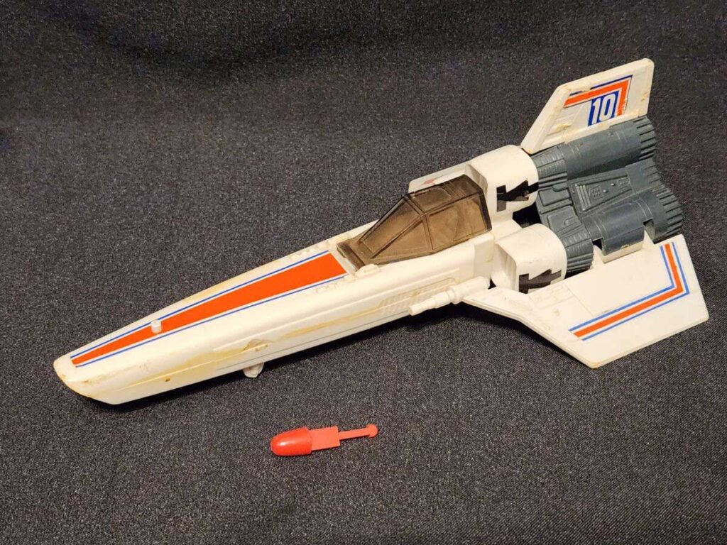 A vintage toy spaceship with a white body, orange and blue stripes, and the number 10 on its wings. The spacecraft is equipped with dual engines at the back, and a small red projectile is positioned below it on a dark fabric surface.