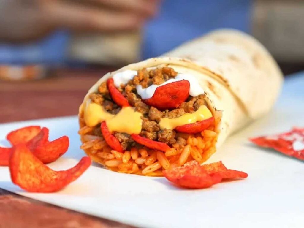 A delicious wrap reminiscent of discontinued Taco Bell menu items, filled with seasoned rice, ground beef, cheese sauce, and jalapeño-flavored chips. Topped with a dollop of sour cream, it rests on a white surface with some chips scattered around.