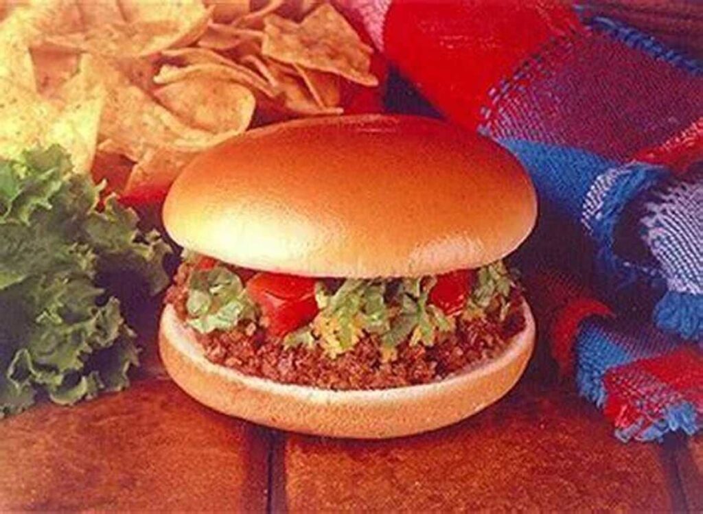 A burger with seasoned beef, chopped lettuce, and tomato on a bun sits next to a pile of chips and leafy greens, reminiscent of beloved discontinued Taco Bell menu items. A colorful cloth lies beside the wooden surface.