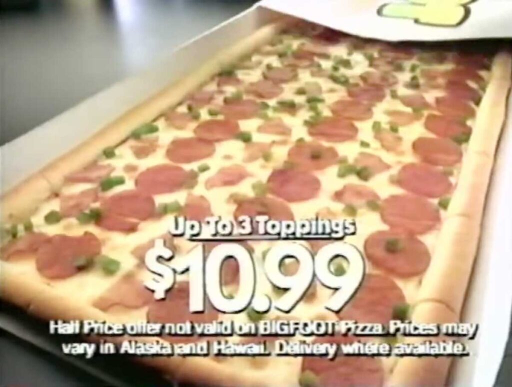 Discontinued fast food item - Pizza Hut's Big Foot Pizza. A rectangular pepperoni and green pepper pizza in a box, with text advertising Up to 3 Toppings $10.99. Small print notes exclusions for discontinued fast food items like BIGFOOT Pizza, and price variations in Alaska and Hawaii. Delivery is mentioned.