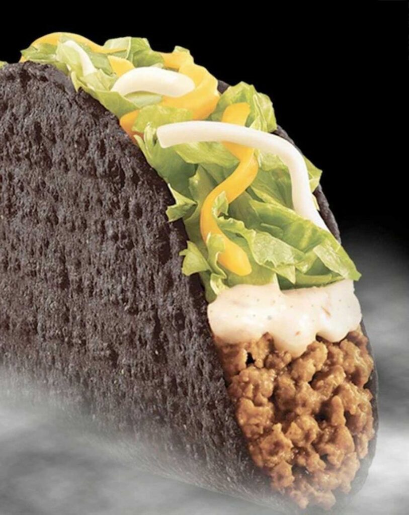 A taco with a black shell, reminiscent of discontinued Taco Bell menu items, filled with seasoned ground beef, shredded lettuce, and a blend of yellow and white cheese, topped with creamy sauce. The background is dark with a misty effect.