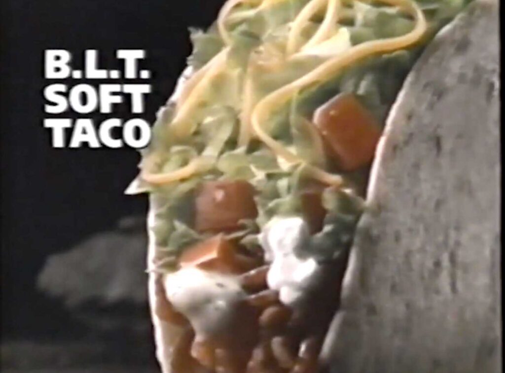 A close-up of a discontinued Taco Bell menu item, the B.L.T. soft taco, is shown with visible shredded lettuce, diced tomatoes, cheese, bacon bits, and creamy sauce inside a soft tortilla. The text on the left reads B.L.T. SOFT TACO.