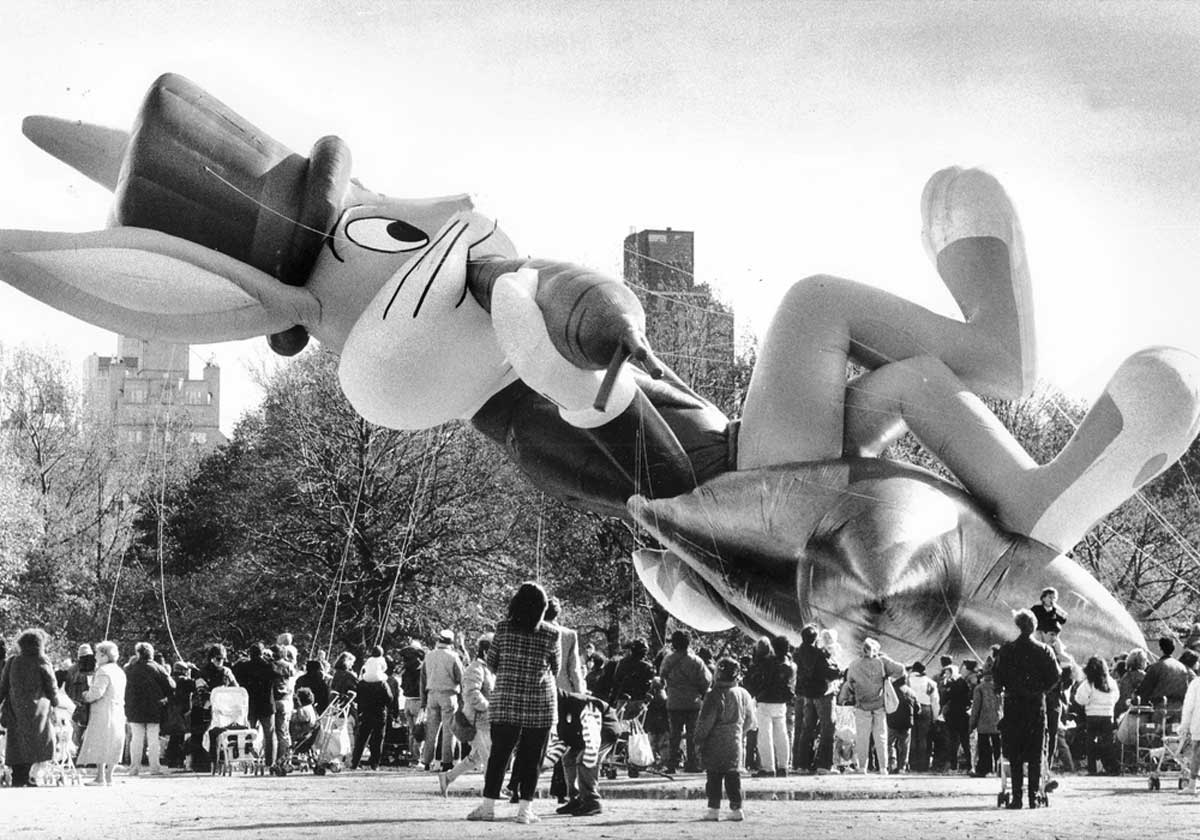 A large cartoon rabbit balloon wearing a top hat is held aloft by ropes during a parade. Crowds gather in a park to watch the festive display, with trees and buildings in the background. The image is in black and white.