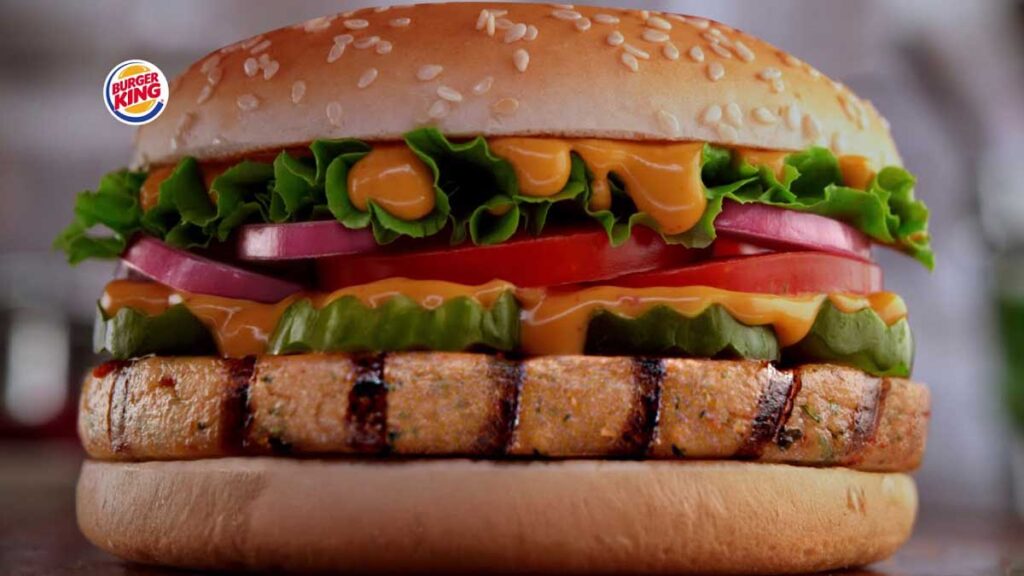 Close-up of a Burger King veggie burger with a sesame seed bun, lettuce, tomato, onions, pickles, and sauce. The patty is grilled to perfection with distinct grill marks. The Burger King logo is visible in the top left corner—a nod to discontinued fast food items that fans still crave.