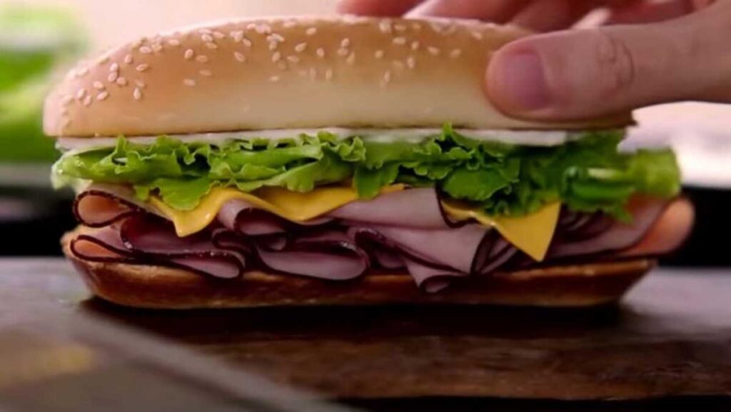 A hand holding a sub sandwich with a sesame seed bun, filled with layers of deli meat, cheese slices, lettuce, and a hint of mayonnaise—a delicious reminder of classic flavors reminiscent of discontinued fast food items.