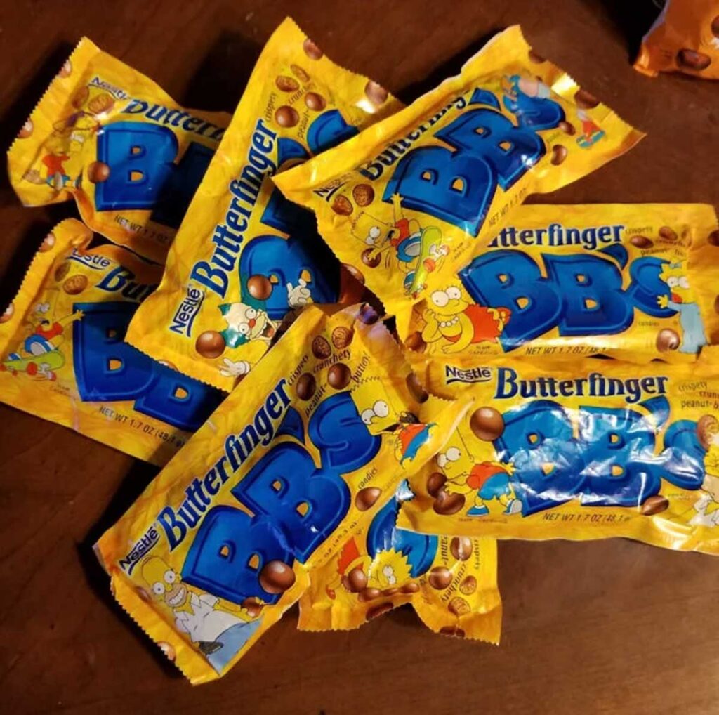 A pile of yellow Butterfinger BBs candy packages, one of the iconic discontinued snacks from the 90s, is spread across a wooden surface. Each package features colorful graphics of cartoon characters and the candy's logo.