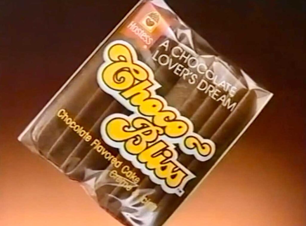 A package of Hostess Choco-Bliss, a beloved yet discontinued snack from the 90s, is displayed with "A Chocolate Lover's Dream" and "Chocolate Flavored Cake" elegantly printed. The background transitions beautifully from dark to light brown.