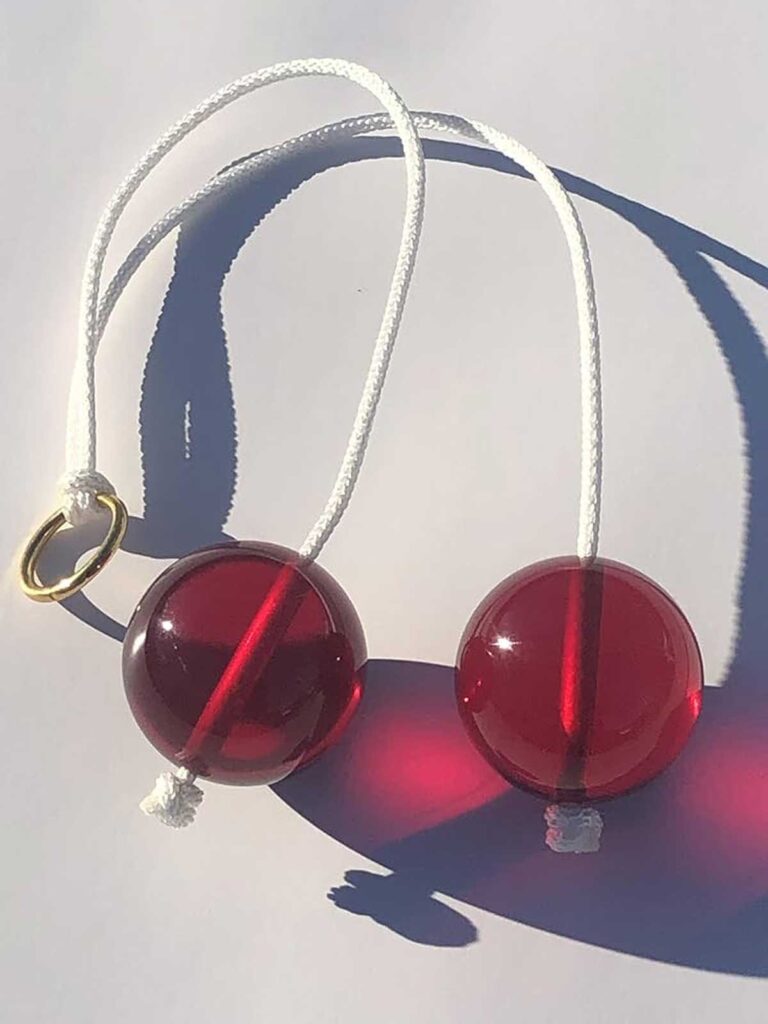 A pair of red acrylic balls attached by a white cord lies on a surface, casting shadows. A gold ring is looped through the cord.