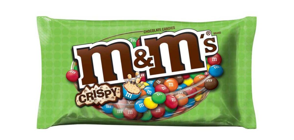 A green bag of crispy M&Ms, echoing the nostalgic charm of discontinued snacks from the 90s, features the brand logo in bold letters. The packaging displays colorful crispy chocolate candies scattered inside a transparent bowl, highlighting their crunchy texture.