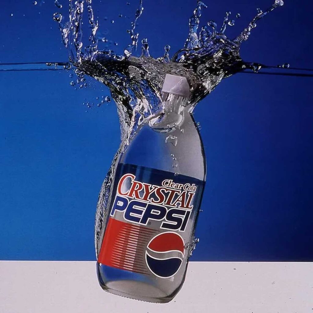 A bottle of Crystal Pepsi plunges into clear water with a splash, reminiscent of beloved, discontinued snacks from the 90s. The label reads Clear Cola Crystal Pepsi and features the iconic red, white, and blue logo against a background divided into blue and white sections.