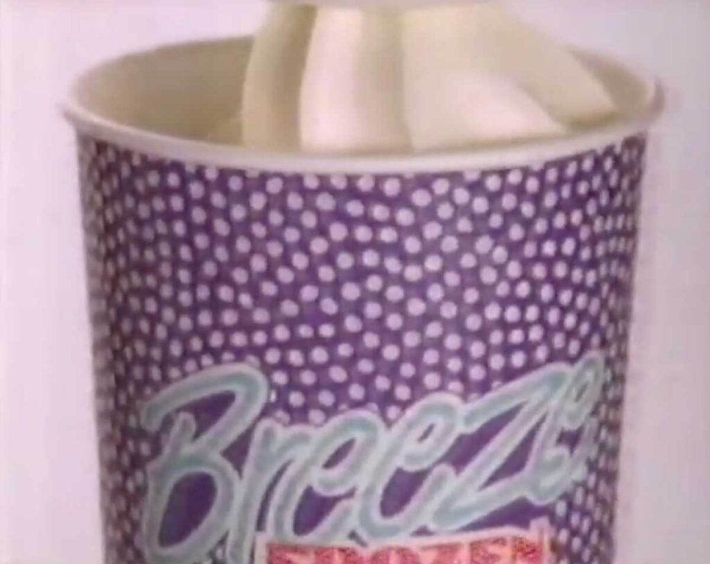 A close-up of a cup filled with soft-serve ice cream evokes nostalgia for discontinued fast food items. The cup is purple with white dots and features the word Breeze in light blue letters, reminiscent of past indulgent delights.