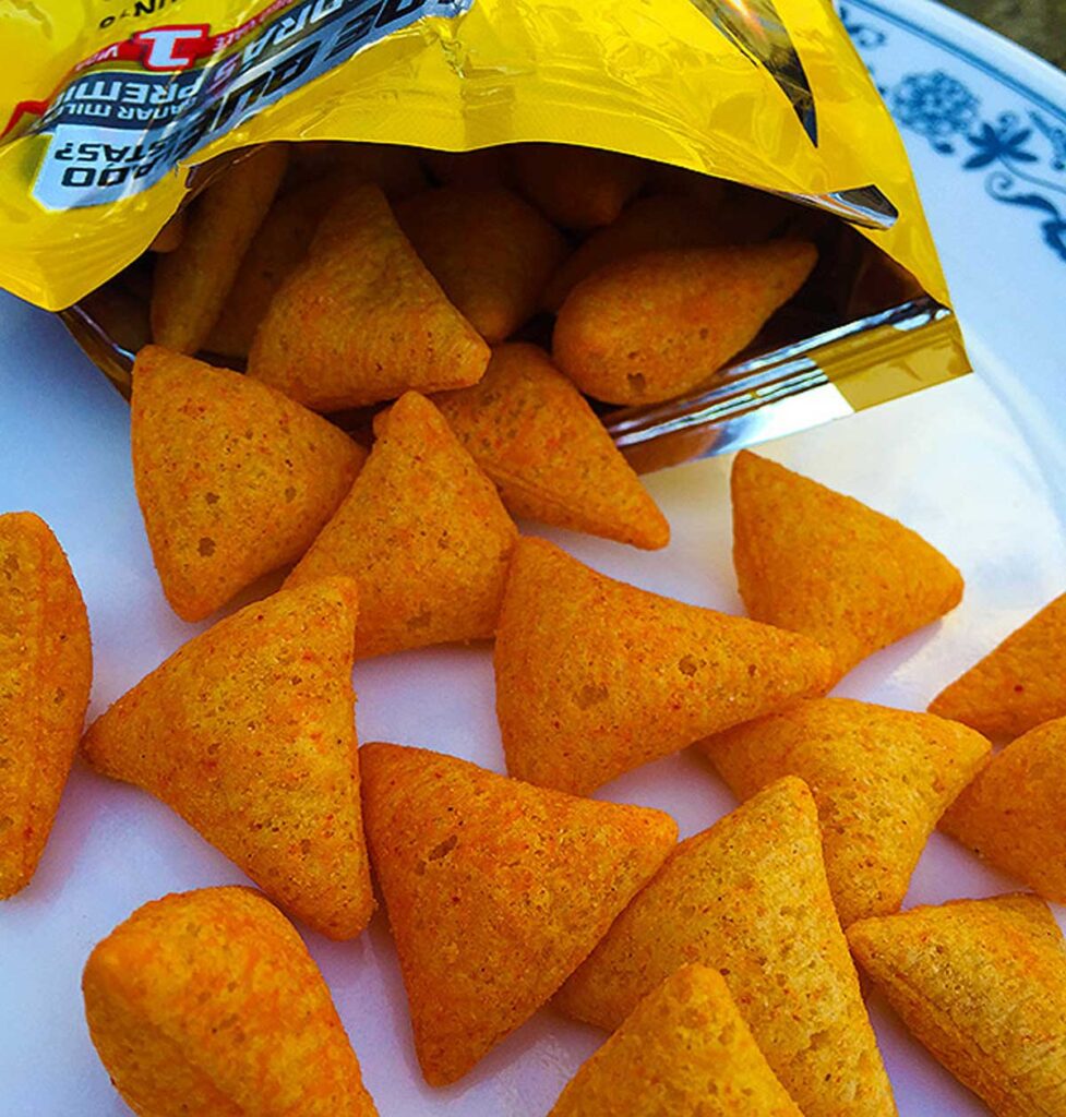 A yellow snack bag with triangular cheese-flavored chips, reminiscent of discontinued snacks from the 90s, spills onto a white plate with a blue floral design. The chips are bright orange and boast a satisfyingly crunchy texture.