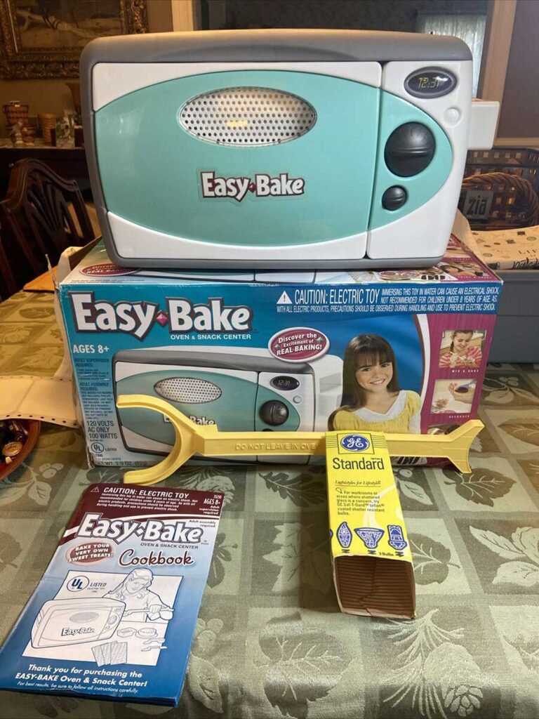 A teal and white Easy-Bake Oven is displayed on top of its box with packaging. Accompanying items include a yellow oven spatula, a package of standard light bulbs, and an Easy-Bake cookbook. The table is covered with a green tablecloth.