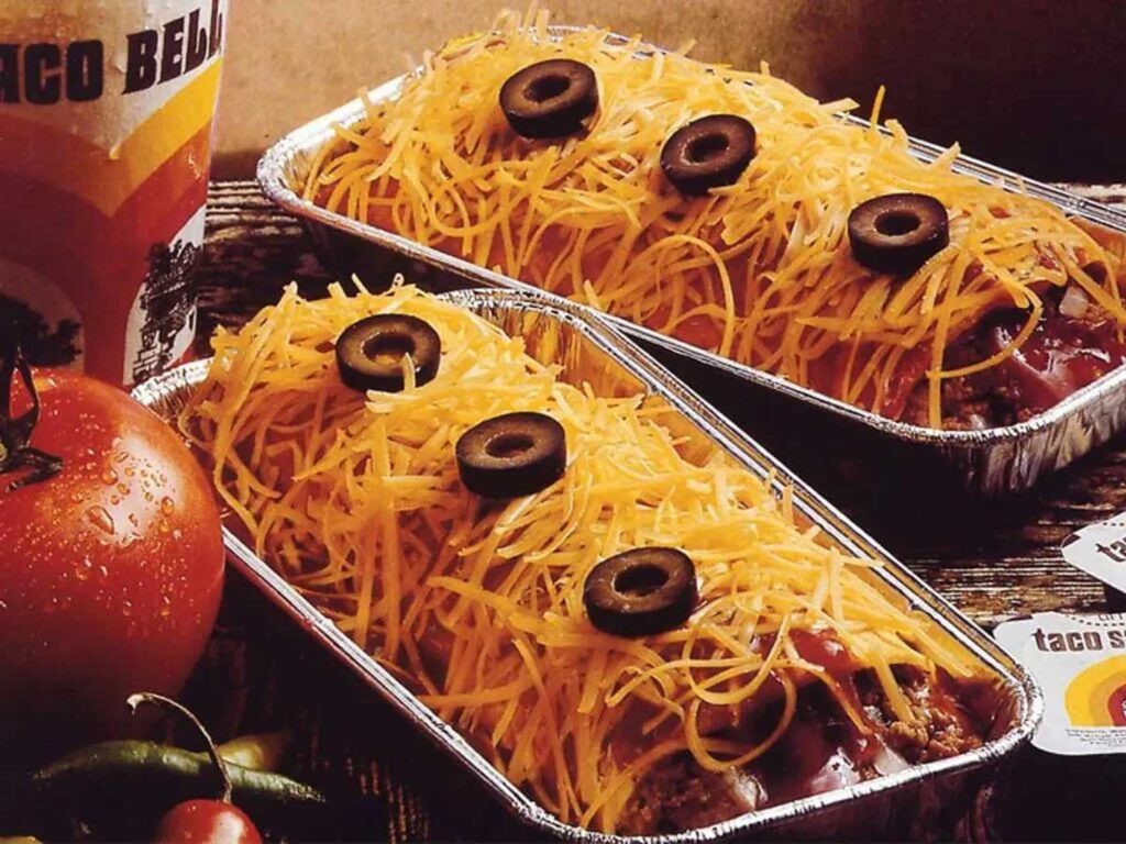 Discontinued Taco Bell Menu Items - Enchirito. Two metal trays filled with enchiladas topped with shredded cheese and sliced black olives evoke memories of beloved, yet discontinued Taco Bell menu items. A juicy tomato and spicy peppers rest on the side, accompanied by a branded cup, against a rustic wooden backdrop.