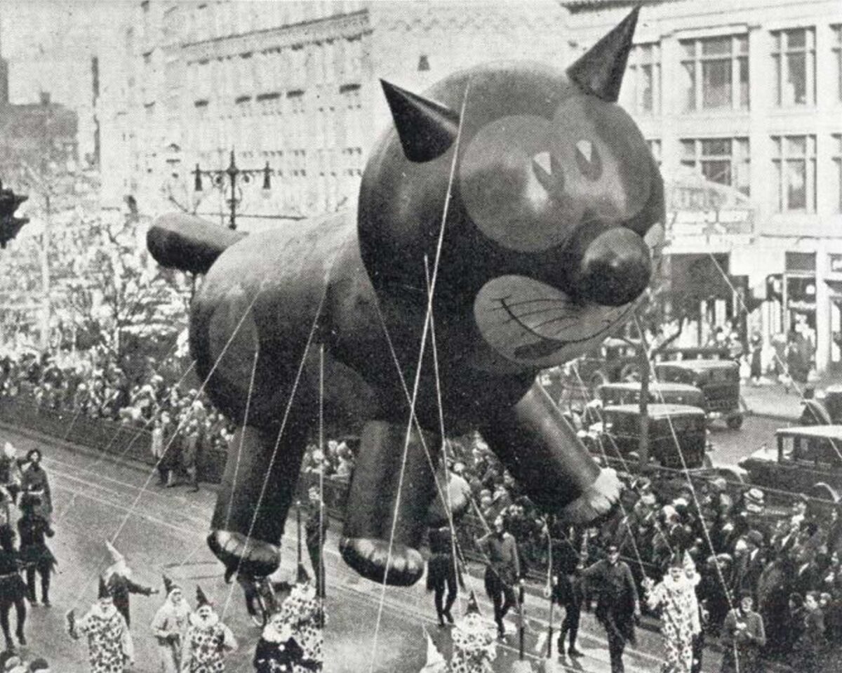 Macy's Thanksgiving Day Parade Fails - A giant balloon of a black cat is being paraded down a city street. A crowd of people lines the street, some holding ropes to guide the balloon. Buildings and vintage cars are visible in the background.