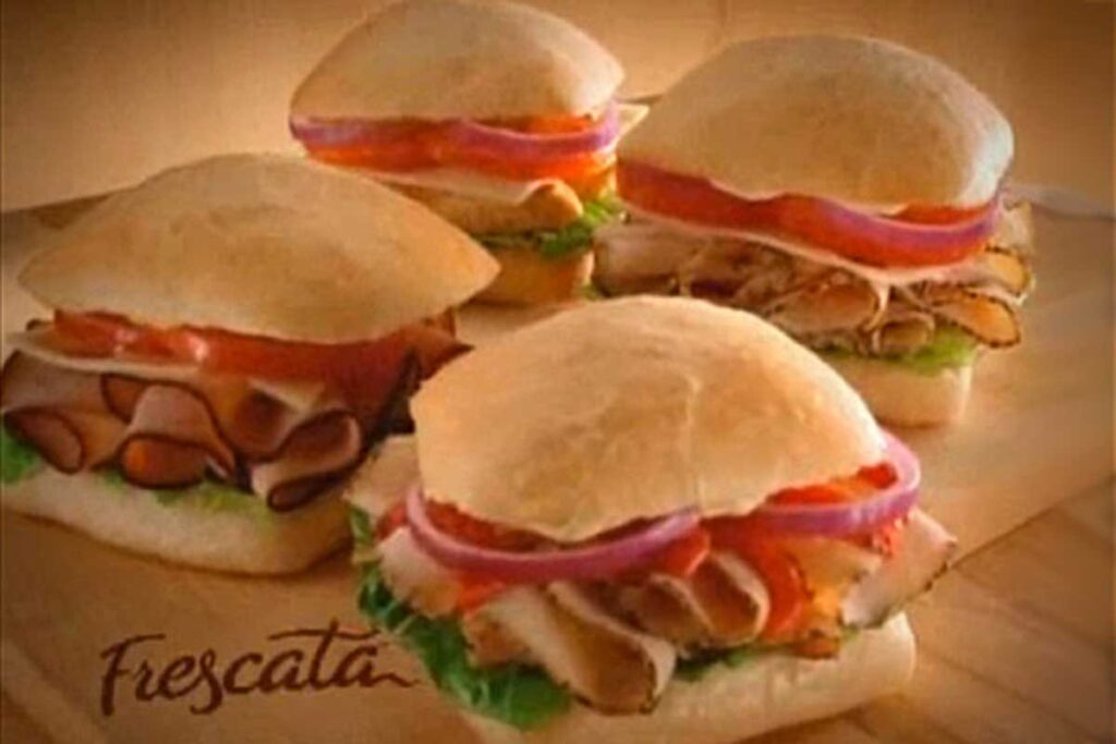 Four sandwiches rest on a wooden surface, each with ciabatta bread. Visible ingredients include slices of tomato, red onion, lettuce, and various deli meats. The word Frescata—evoking memories of beloved discontinued fast food items—graces the bottom left corner.