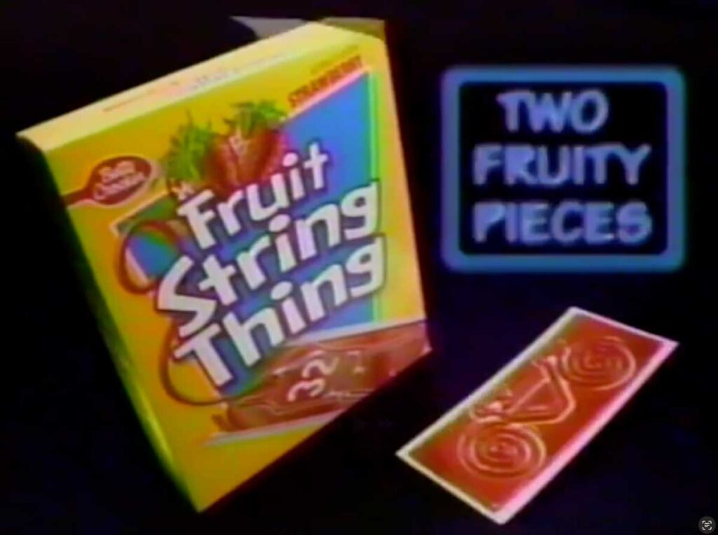 A box of Betty Crocker Fruit String Thing in strawberry flavor is displayed, a nostalgic nod to discontinued snacks from the 90s. The box features colorful graphics, and next to it, two rolled fruit strings await. A sign reads Two Fruity Pieces.