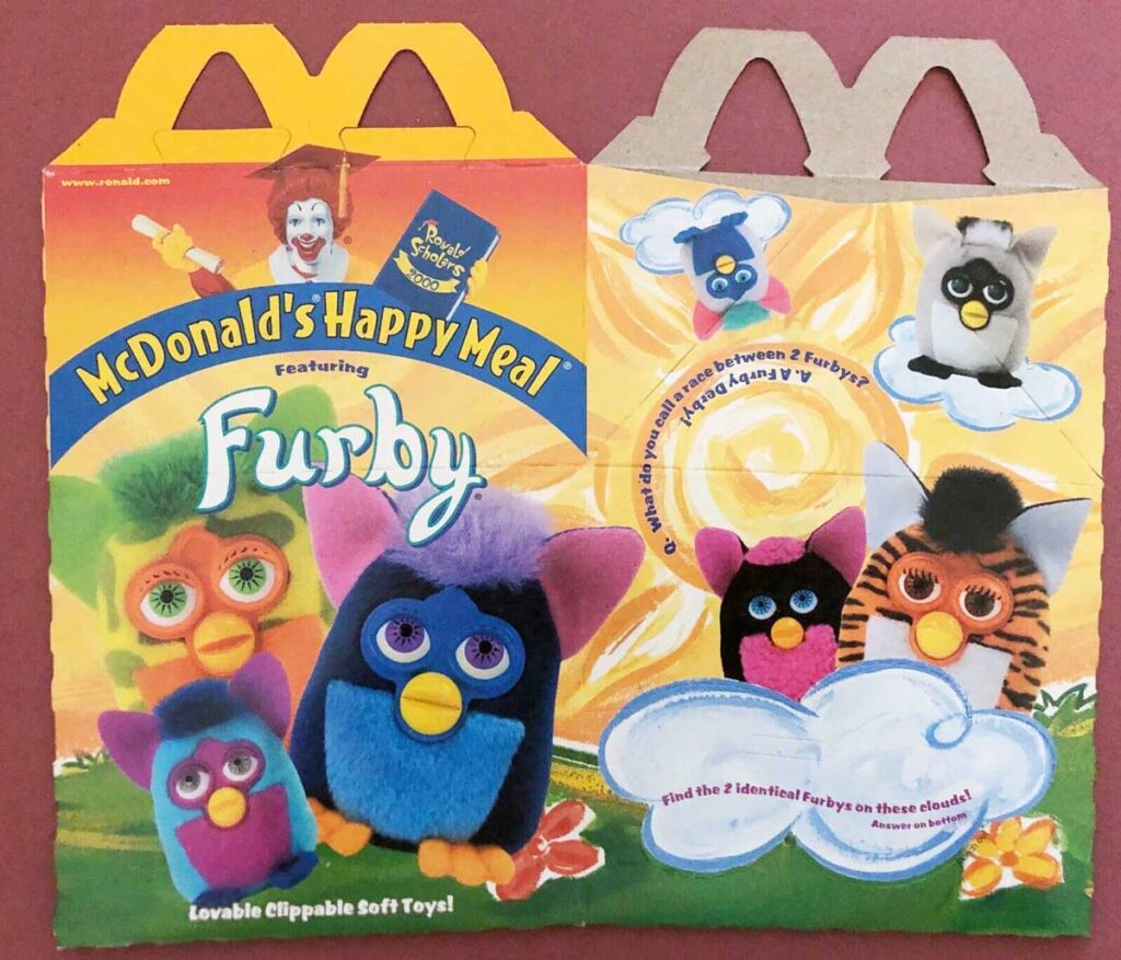 The image shows a colorful McDonalds Happy Meal box featuring Furbys. It includes playful, fluffy Furby toy illustrations in vibrant colors, encouraging kids to find two identical Furbys among clouds. The design is bright and eye-catching.