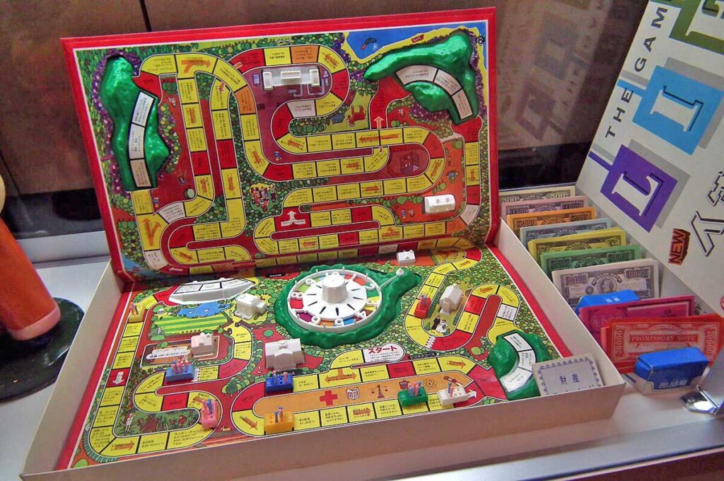 A colorful board game with winding paths and various spaces. Several miniature cars, houses, and pegs are positioned on the board. Play money and cards are organized to the side. The game is set up on a tabletop.