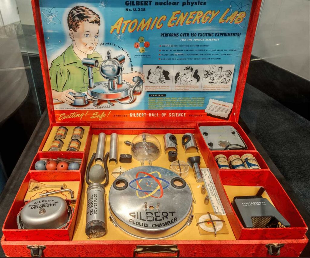 Vintage Atomic Energy Lab science kit in an open red case. Includes tools like a Geiger counter, cloud chamber, radioactive samples, and instruction booklet, with colorful packaging featuring a child scientist illustration on the lid.