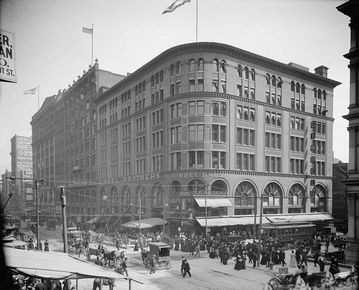 A black and white photo captures a bustling city street corner during Gimbel's Thanksgiving Day Parade, featuring a large, multi-story building with an arched facade and a sign reading Siegel Brothers. Horse-drawn carriages and excited pedestrians fill the lively street.