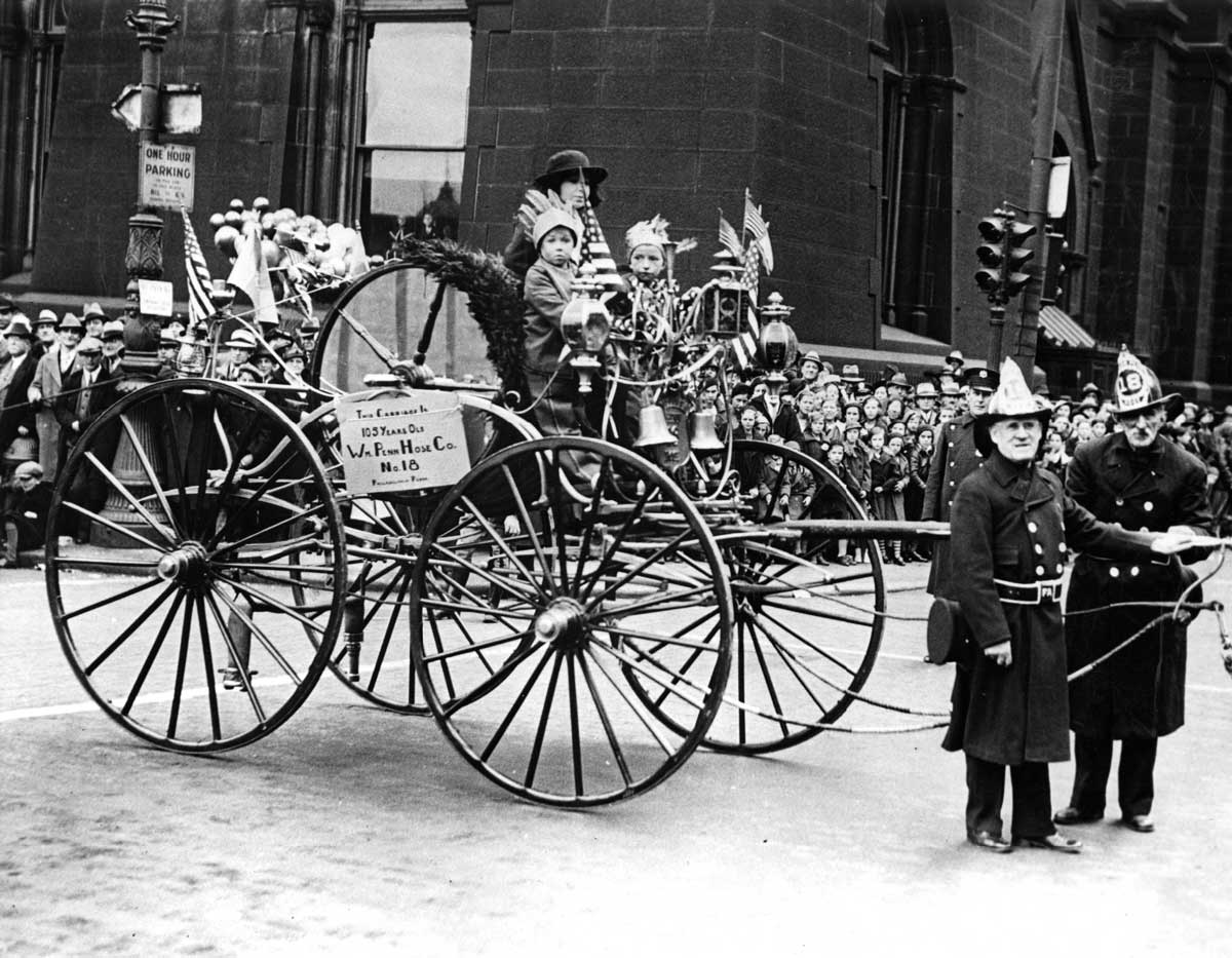 A vintage black and white photo shows a festive parade with a horse-drawn carriage carrying children in costumes. Two firefighters stand in front, while a crowd watches along the street.