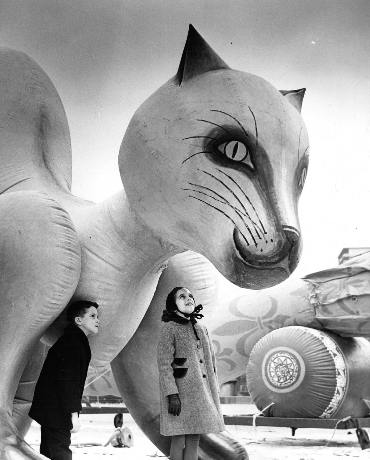 Two children stand beside a giant cat balloon, looking up in awe at the whimsical spectacle. With a cartoon-like expression, it towers over them against a backdrop of cloudy skies, evoking nostalgic scenes from Gimbel's Thanksgiving Day Parade.