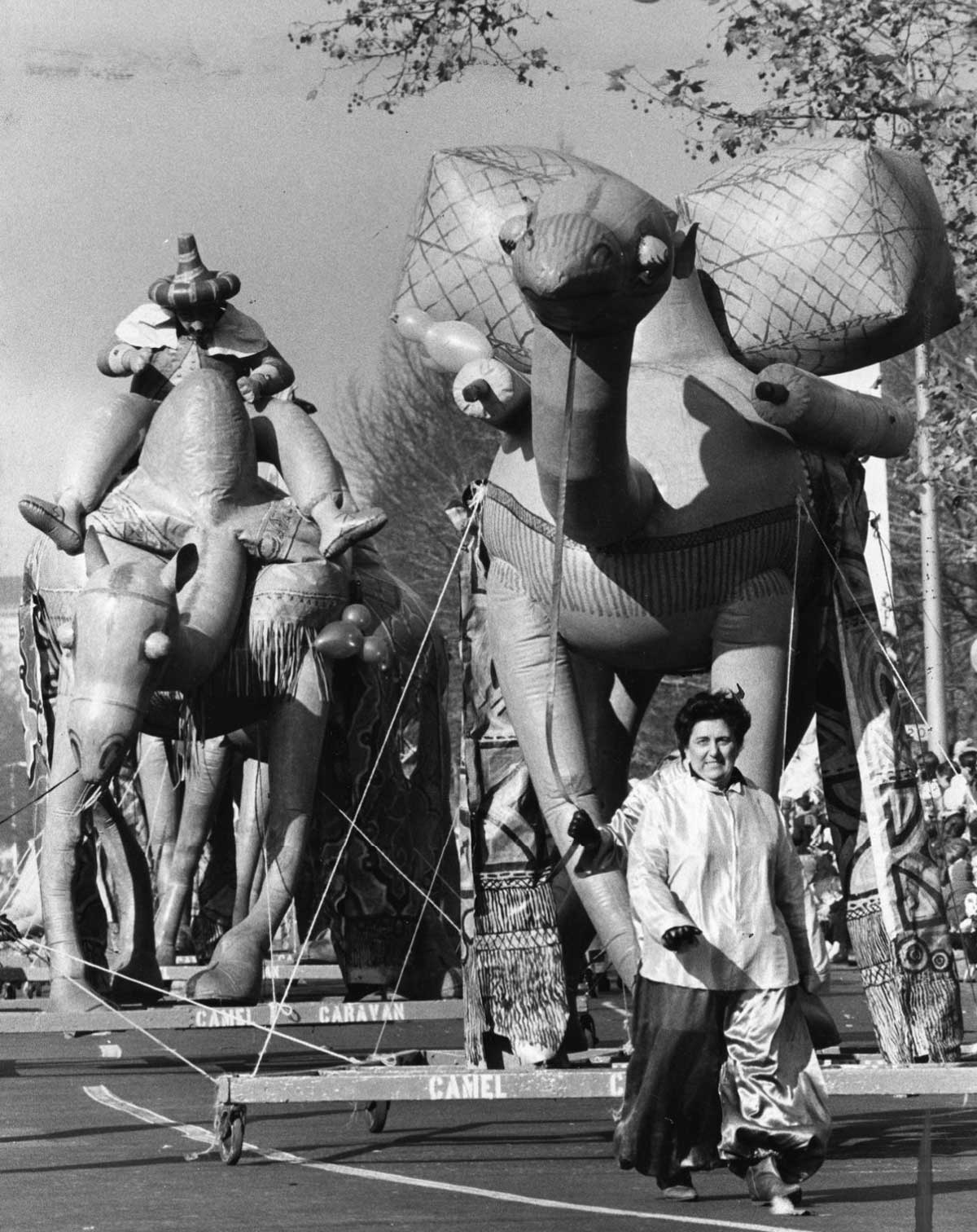 A woman walks beside large inflatable animal balloons shaped like camels and elephants during a street parade. Trees and a crowd of spectators are visible in the background. The scene is lively and festive.