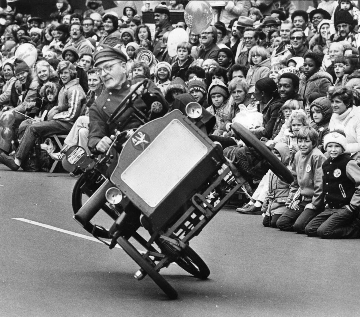 During the Gimbel's Thanksgiving Day Parade, a man in a military-style hat rides a vintage motorized vehicle on two wheels. The excited crowd, brimming with children, watches in amusement. The scene is energetic and lively, perfectly capturing the joy of this festive event.