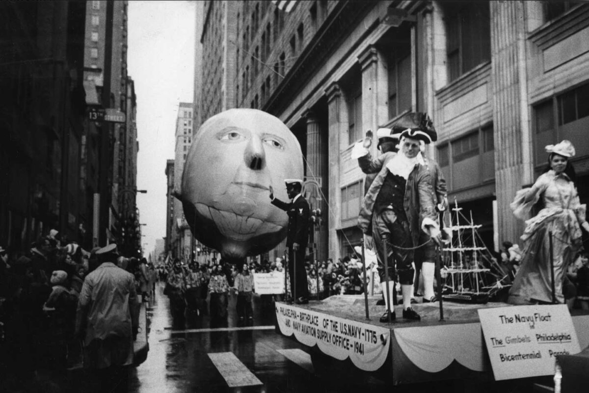 A black and white photo captures the Gimbel's Thanksgiving Day Parade, showcasing a large George Washington head balloon, a float with people in 18th-century costumes, and a sign highlighting The Navy Float at The Gimbels Philadelphia Bicentennial. Enthusiastic crowds line the streets.