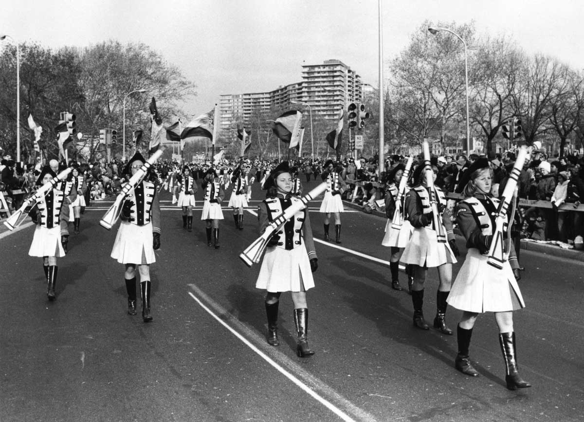 A black and white photo of a marching band parade. Uniformed individuals in skirts and jackboots march down a wide street holding instruments. Spectators line the street, flanked by tall buildings and leafless trees.