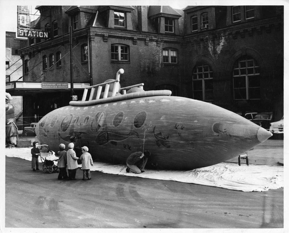 A vintage black and white photo of people standing around a large, detailed model of a submarine on a city street. The submarine is situated in front of a historic building with a Station sign. Several children and adults observe the display.