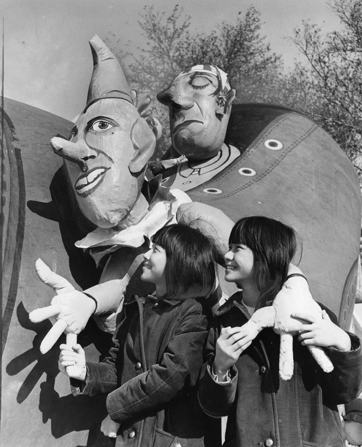 Two children gaze up at a large carnival float featuring whimsical, oversized faces of clowns, with exaggerated features. The float is adorned with artistic details, and it is set against a backdrop of trees and a clear sky.