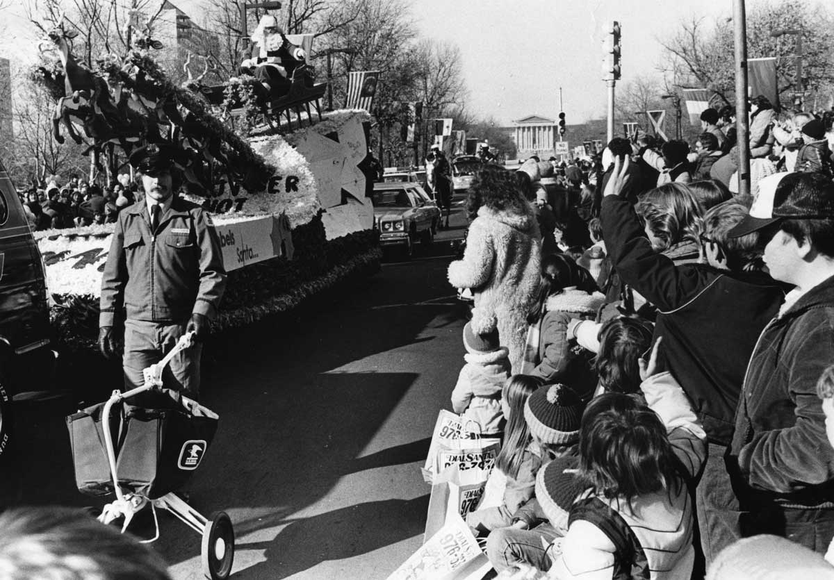 A black and white photo of a parade featuring a float with Santa and reindeer. A large crowd lines the street, with people, including children, waving and holding signs. A person in uniform walks alongside the float.