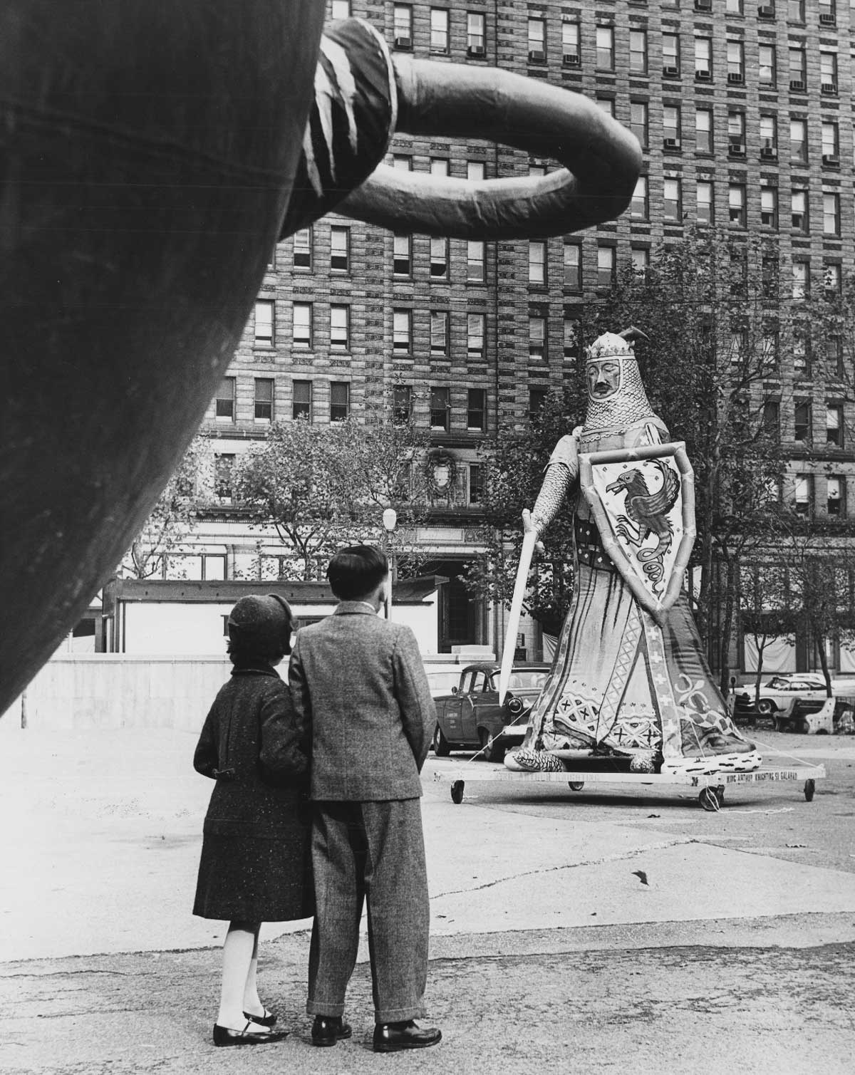 A boy and girl stand on a city street, mesmerized by the towering inflatable knight with a sword and shield, reminiscent of the magic found at Gimbel's Thanksgiving Day Parade. Behind them looms a tall building, completing the enchanting urban scene.