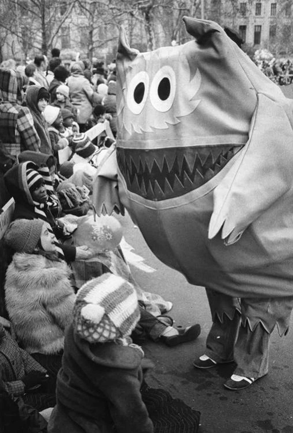 A child-friendly parade with a person in a large monster costume interacting with smiling children seated along the sidewalk. The monster has big eyes, pointy teeth, and horns. The scene is festive with children wearing winter clothes.