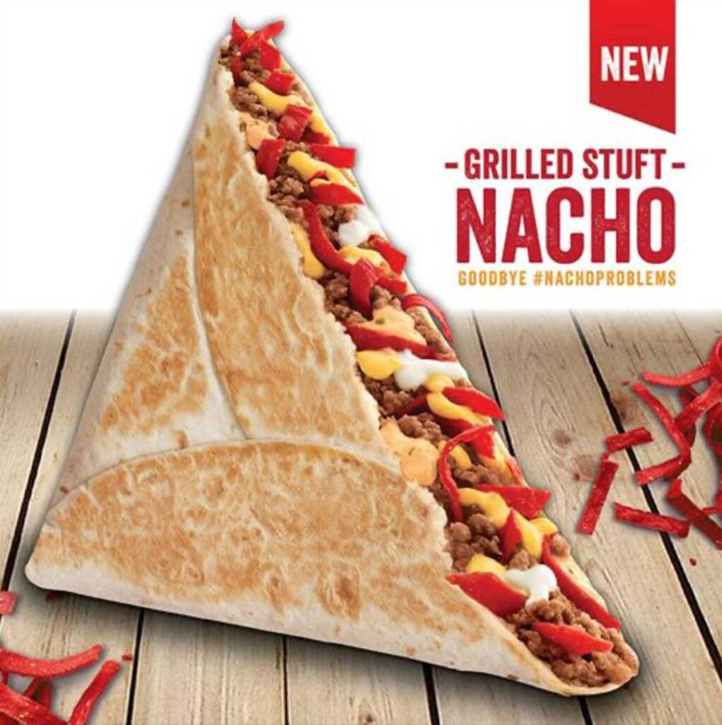 A triangular grilled stuffed nacho filled with seasoned meat, cheese, and red strips sits on a wooden surface. Red strips and text reading New Grilled Stuft Nacho appear in the background—a delicious throwback to beloved discontinued fast food items.