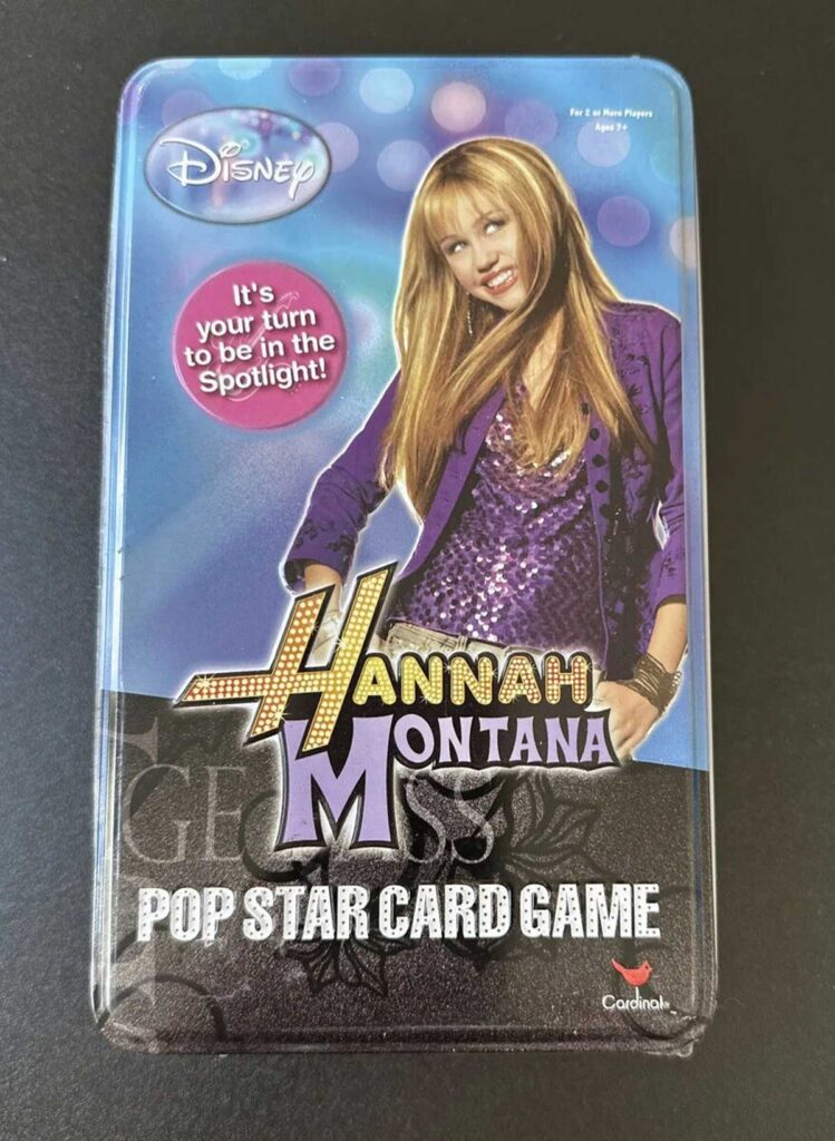 A Disney Hannah Montana Pop Star Card Game tin featuring a smiling girl with long blonde hair, wearing a glittery purple outfit against a blue background. Text bubble says, Its your turn to be in the spotlight!.