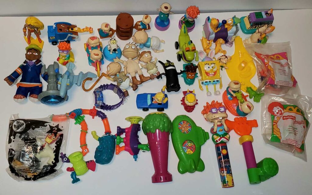 A collection of colorful vintage toys and figurines from various animations is spread out on a white surface. The assortment includes characters, vehicles, and packaging with vibrant colors and diverse shapes.