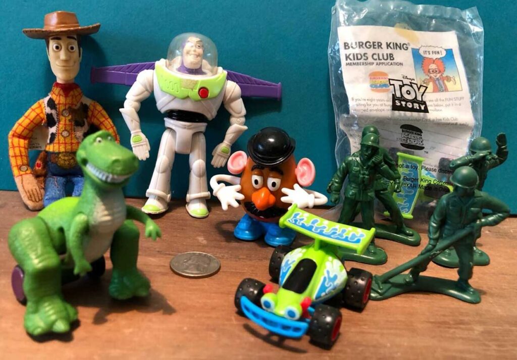 Toy Story figurines, including a cowboy, astronaut, dinosaur, potato head, and green soldiers, are arranged around a small toy car. A Burger King Kids Club package is in the background.