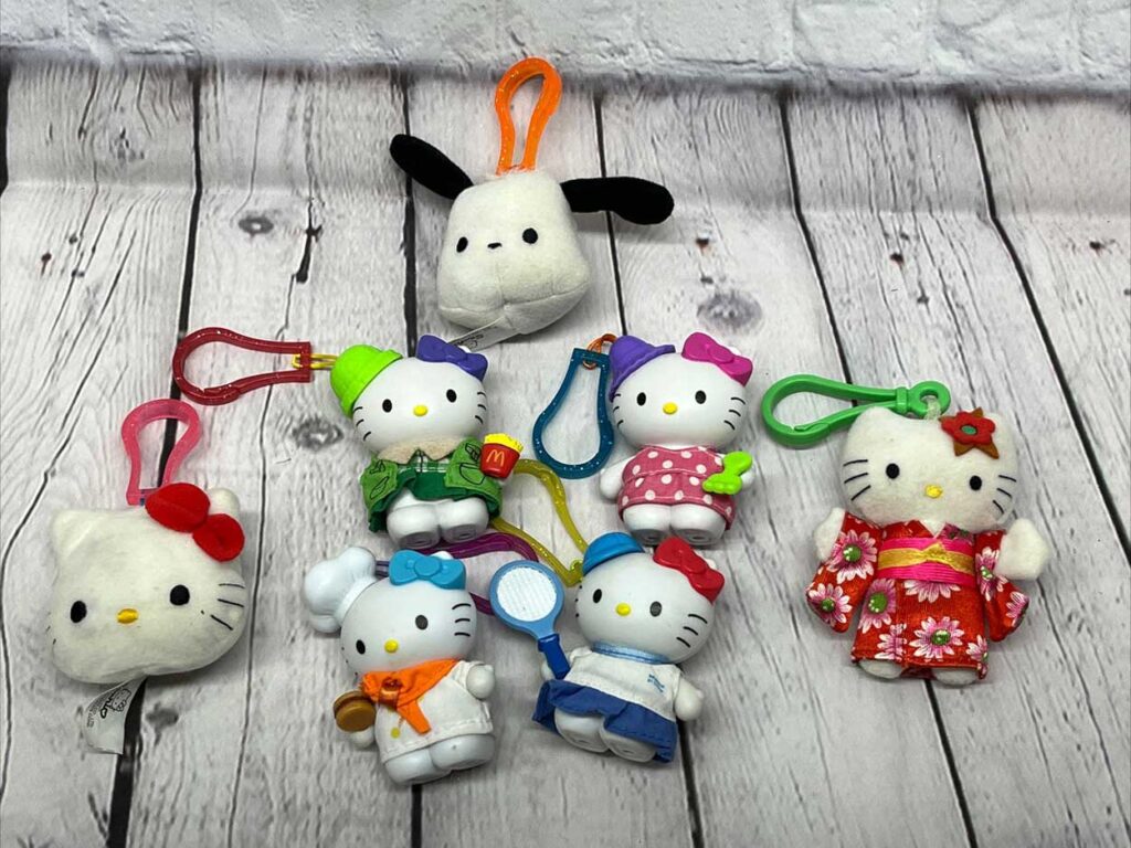 A collection of seven Hello Kitty and animal-themed keychains displayed on a wooden surface. They feature bright colors and various outfits, including a red kimono, a chefs outfit, and polka dots.