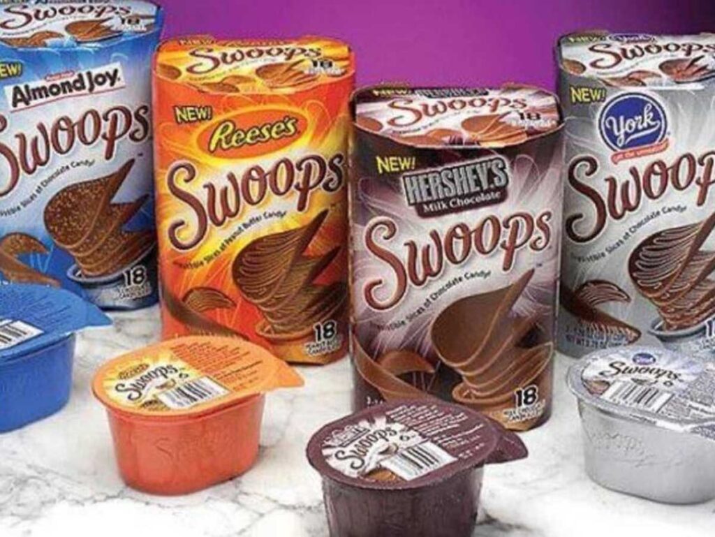 A variety of Swoops candy packs, reminiscent of discontinued snacks from the 90s, are displayed on a purple and white surface. Flavors include Almond Joy, Reeses, Hersheys Milk Chocolate, and York. Each box contains 18 pieces with individual containers in front.