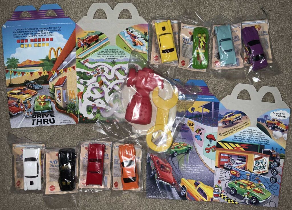 McDonalds toy car set laid out on a carpet. Includes seven toy cars in packaging and two large red and yellow plastic tools, displayed with colorful cardboard backdrops featuring drive-thru illustrations.