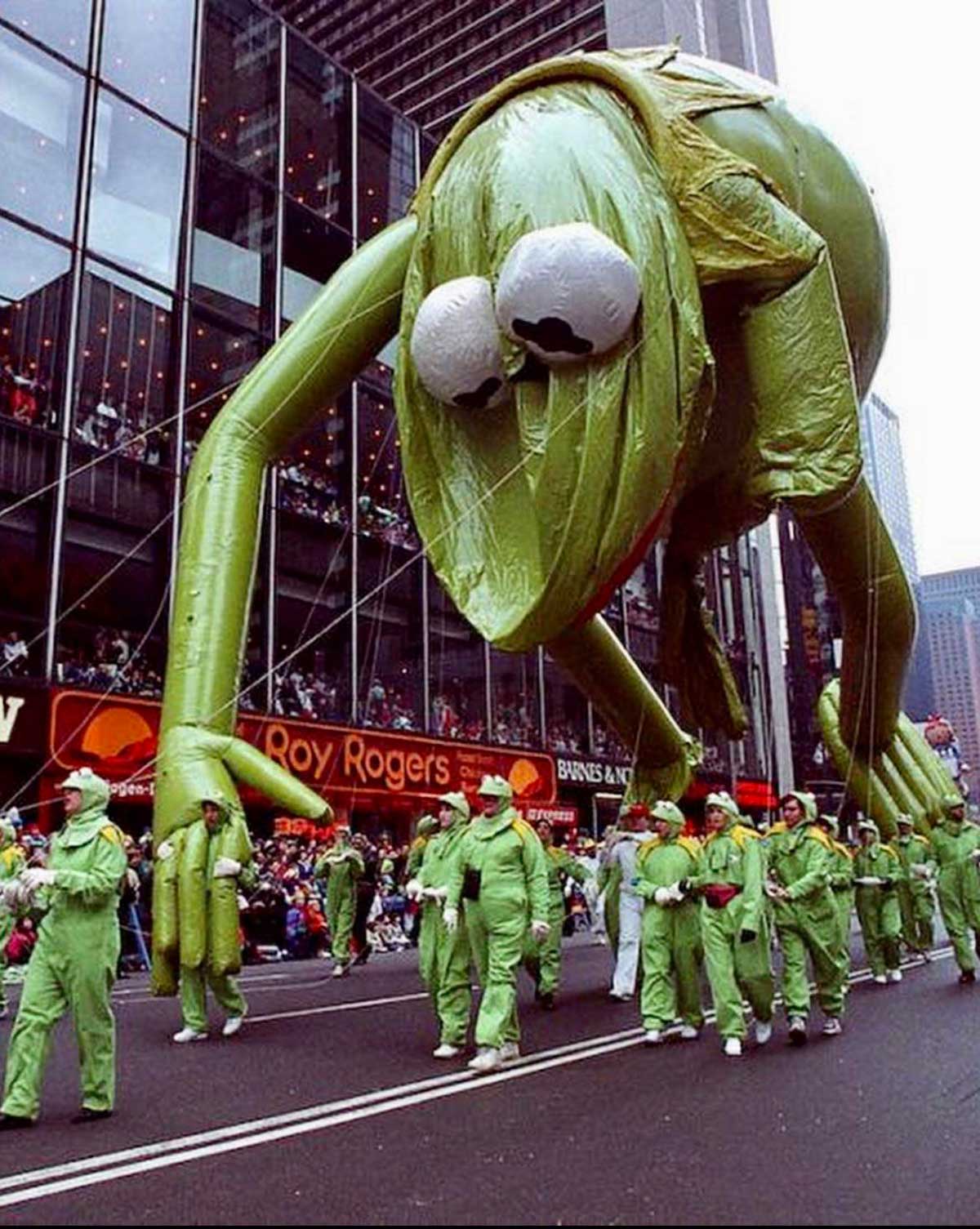 A large green frog balloon with big eyes floats above a street during a parade. People in matching green costumes walk below, guiding the balloon. Spectators watch from the sidewalks and a nearby building.